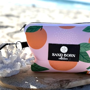 Op de afbeelding: Een tasje met rits en een lichtroze achtergrond met een patroon van sinaasappels, groene bladeren en witte bloemen. Een zwart rechthoekig label op de voorzijde toont de tekst SAND BORN collection met een zanddollar-icoon. Een zwarte polsband is bevestigd.