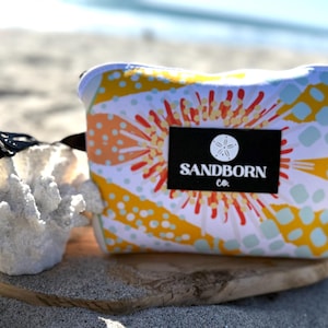 May include: A rectangular pouch featuring a vibrant pattern of yellow, orange, and light blue abstract shapes and dots. A black rectangular label is centered on the front, displaying a white sand dollar icon and the text SANDBORN CO.
