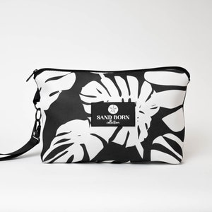 May include: Black and white zippered pouch with a tropical leaf print and a black strap. The pouch has a logo that says "SAND BORN collection".