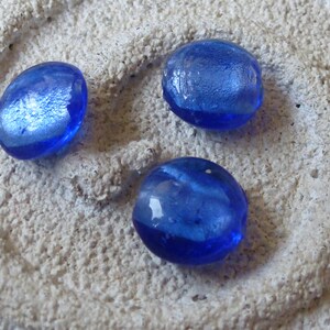 Könnte beinhalten: Drei blaue Glasperlen mit einem metallischen Schimmer. Die Perlen sind oval geformt und haben eine glatte, glänzende Oberfläche.