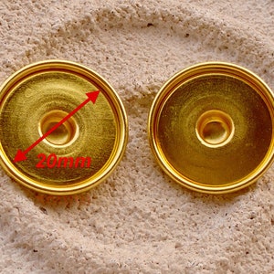 Könnte beinhalten: Zwei goldfarbene Metall-Druckknöpfe mit einem Durchmesser von jeweils 20 mm. Die Knöpfe haben eine glatte, glänzende Oberfläche und einen erhöhten Rand.