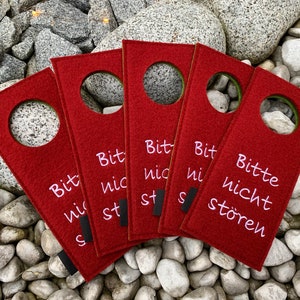 May include: Five red felt door hangers with a white circle in the center and the text "Bitte nicht st&ouml;ren" in pink lettering. The text translates to "Please do not disturb".