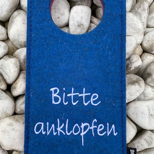 May include: A blue felt door hanger with a round hole in the center and the text "Bitte anklopfen" in white embroidery. The bottom right corner has a small white logo with the text "Filzlaus".