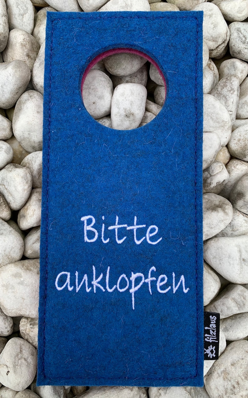 May include: A blue felt door hanger with a round hole in the center and the text "Bitte anklopfen" in white embroidery. The bottom right corner has a small white logo with the text "Filzlaus".