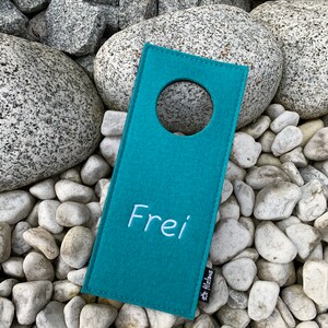 May include: Teal felt door hanger with a round hole and the word "Frei" embroidered in white. The hanger has a small white tag with the word "Filzinas" on it.