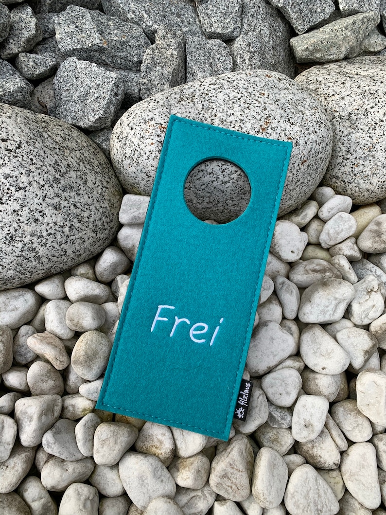 May include: Teal felt door hanger with a round hole and the word "Frei" embroidered in white. The hanger has a small white tag with the word "Filzinas" on it.
