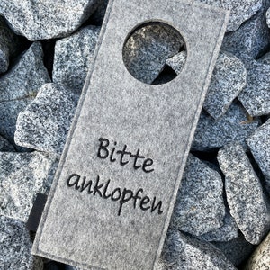 May include: Gray felt door hanger with a round cutout and the text "Bitte anklopfen" in black embroidery.