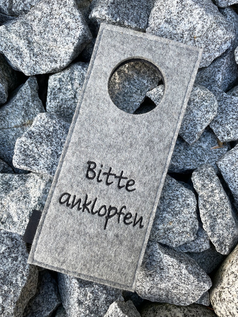 May include: Gray felt door hanger with a round cutout and the text "Bitte anklopfen" in black embroidery.