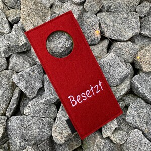 May include: Red felt door hanger with a round hole in the center and the word "Besetzt" embroidered in white.