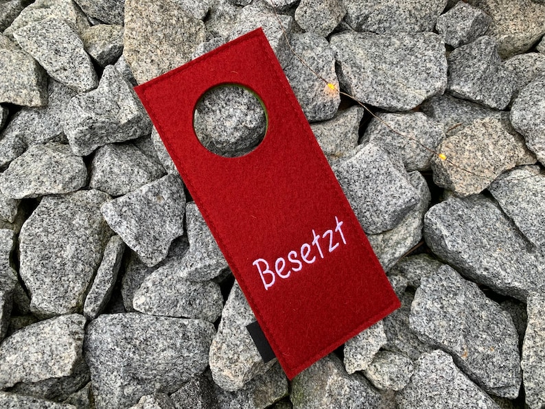 May include: Red felt door hanger with a round hole in the center and the word "Besetzt" embroidered in white.