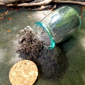 May include: A glass jar with a cork lid, filled with a dark gray powder. The jar is tipped over, spilling the powder onto a gray surface.