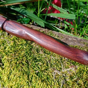 May include: A handcrafted, dark reddish-brown wooden object with a hole at one end. The wood grain is visible, and the object has a smooth, polished finish. It rests on a bed of green moss, with hints of grass and foliage in the background.