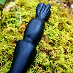 May include: A black wooden wand with a pointed tip and a textured, brush-like top. The wand is resting on a bed of green moss.