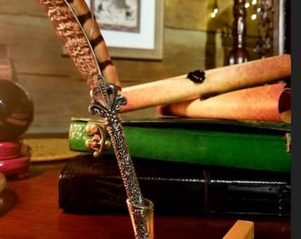 May include: A silver quill pen with a brown and black feather sits in a silver stand on a wooden desk. A small silver letter opener and a stick of sealing wax are also on the desk.