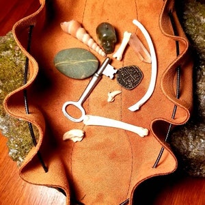 May include: A brown leather pouch with a drawstring closure. The pouch is filled with various items, including a key, a stone, a seashell, a bone, and a small metal charm.
