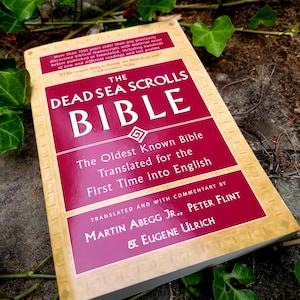 May include: A red and gold book titled "The Dead Sea Scrolls Bible: The Oldest Known Bible Translated for the First Time into English" by Martin Abegg Jr., Peter Flint & Eugene Ulrich.