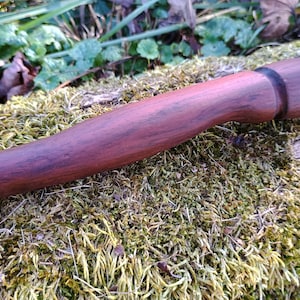 May include: A handcrafted wooden object with a rich, dark brown finish. The object has a cylindrical shape with a slightly curved handle and a wider, rounded end. The wood grain is visible, adding to its natural aesthetic. The object rests on a bed of green moss.