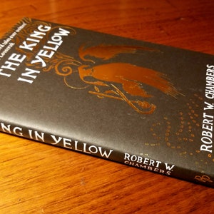 May include: A black hardcover book titled "The King in Yellow" by Robert W. Chambers. The book cover features a gold illustration of a figure with wings and a crown.