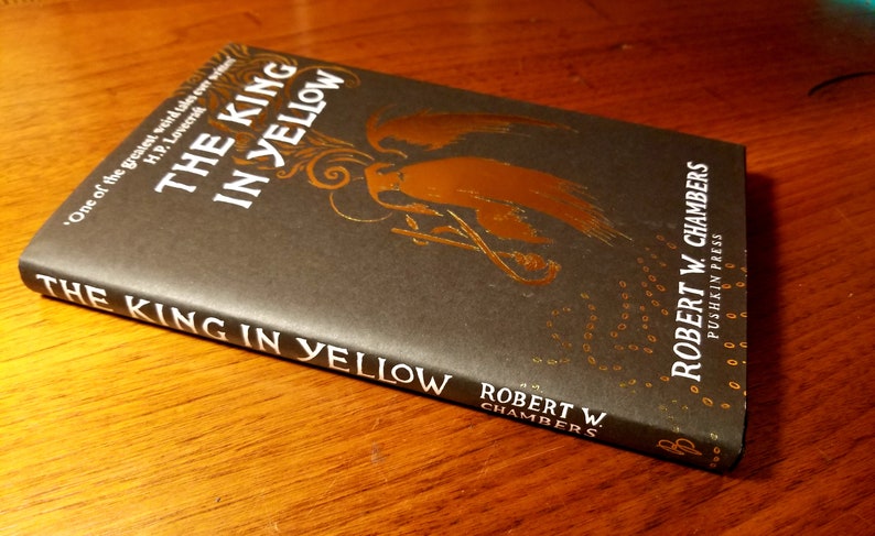 May include: A black hardcover book titled "The King in Yellow" by Robert W. Chambers. The book cover features a gold illustration of a figure with wings and a crown.