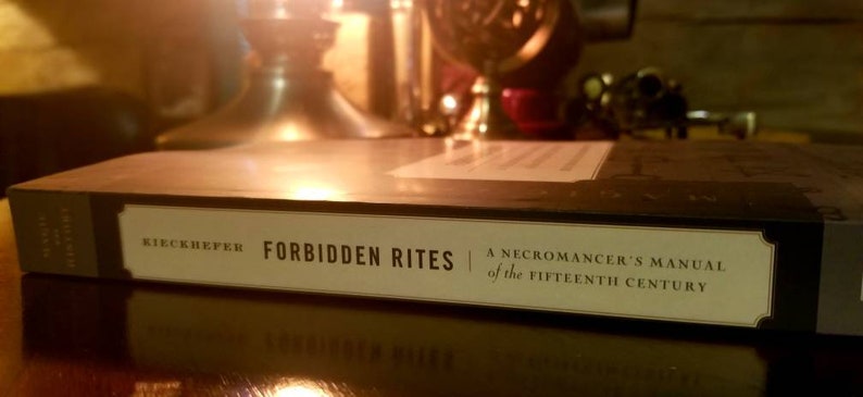 K&ouml;nnte beinhalten: Ein schwarzes Hardcover-Buch mit dem Titel "Verbotene Rituale: Ein Nekromantenhandbuch aus dem 15. Jahrhundert" von Kiechhefer.