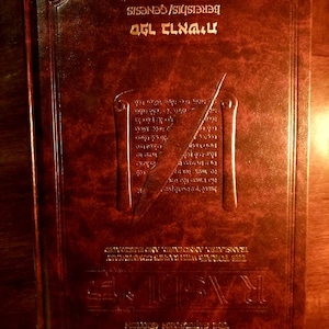 May include: A brown leather-bound book with gold lettering on the cover. The title is "ספר בראשית" which translates to "Genesis" in English. The book is closed and the spine is visible.