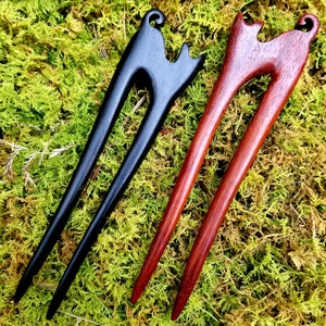 May include: Two wooden hair sticks with cat head designs. One stick is black and the other is reddish brown.