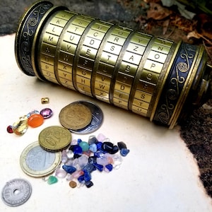 May include: A gold-colored metal cylinder with a combination lock featuring letters of the alphabet. The cylinder is lying on a light brown surface with a collection of colorful gemstones and coins scattered around it.