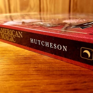 May include: A red hardcover book titled "North American Folk Magic" by Cory Hutcheson. The book has a crescent moon symbol on the spine.