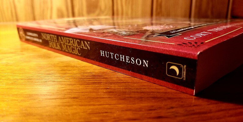 May include: A red hardcover book titled "North American Folk Magic" by Cory Hutcheson. The book has a crescent moon symbol on the spine.