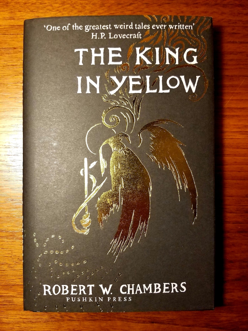 May include: A black book cover with gold lettering that reads "The King in Yellow" by Robert W. Chambers. The cover features a gold illustration of a winged creature with a long, thin tail.