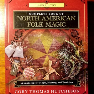May include: A red book with a gold and black title that reads "Llewellyn's Complete Book of North American Folk Magic". The book cover features a landscape of magic, mystery, and tradition with a straw doll, herbs, and other magical objects.