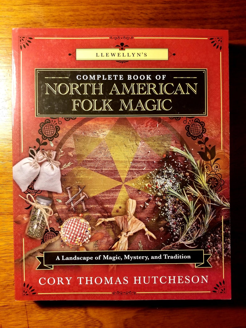 May include: A red book with a gold and black title that reads "Llewellyn's Complete Book of North American Folk Magic". The book cover features a landscape of magic, mystery, and tradition with a straw doll, herbs, and other magical objects.