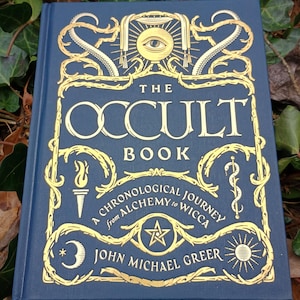 Puede incluir: Un libro de tapa dura azul con letras e ilustraciones doradas. El t&iacute;tulo es "The Occult Book: A Chronological Journey from Alchemy to Wicca" de John Michael Greer.
