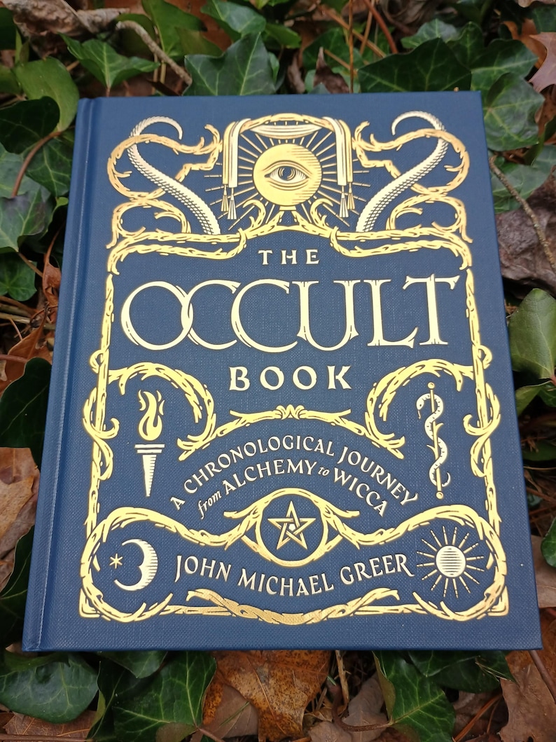 Puede incluir: Un libro de tapa dura azul con letras e ilustraciones doradas. El t&iacute;tulo es "The Occult Book: A Chronological Journey from Alchemy to Wicca" de John Michael Greer.
