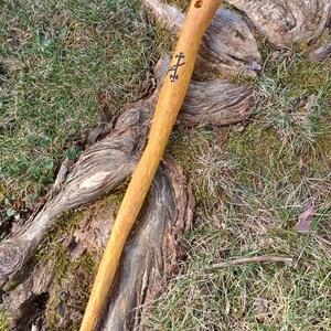 以下が含まれることがあります： 明るい茶色の仕上げが施された、手作りの木製杖。杖には彫刻された十字のデザインと上部の穴があります。杖は草と木の根を背景にしています。