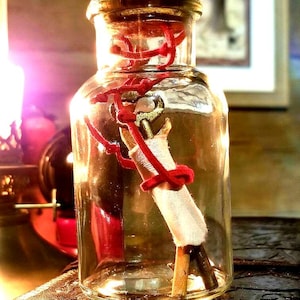 May include: A clear glass bottle with a cork stopper, containing a rolled up piece of paper tied with red string and a small golden key. The bottle is sitting on a brown, textured surface.