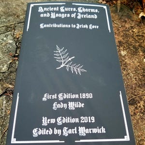 May include: A black book cover with white text that reads "Ancient Cures, Charms, and Usages of Ireland Contributions to Irish Lore First Edition 1890 Lady Wilde New Edition 2019 Edited by Carl Warwick".