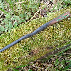 May include: A handcrafted wooden wand with a dark gray handle and a brown top with green vine-like accents. The wand is lying on a bed of green moss and surrounded by grass.