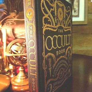 Puede incluir: Un libro negro de tapa dura con letras doradas titulado "The Occult Book: A Chronological Journey Through Alchemy and Witchcraft by John Michael Greer". La portada del libro presenta un ojo con iris y pupila dorados.