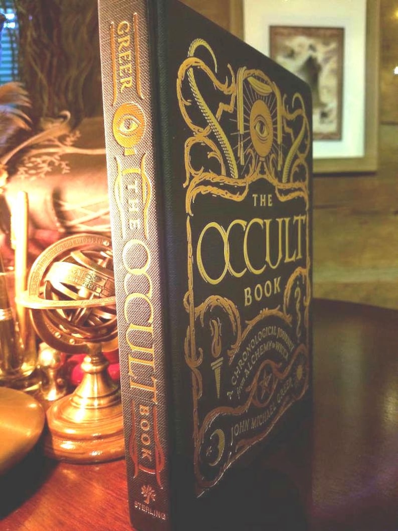 Puede incluir: Un libro negro de tapa dura con letras doradas titulado "The Occult Book: A Chronological Journey Through Alchemy and Witchcraft by John Michael Greer". La portada del libro presenta un ojo con iris y pupila dorados.