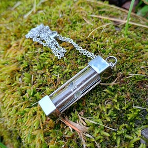 May include: A silver-toned metal hourglass pendant necklace featuring a clear glass cylinder filled with fine dark sand, held within a rectangular frame with a matching silver chain attached to the top loop for wearing as jewelry.