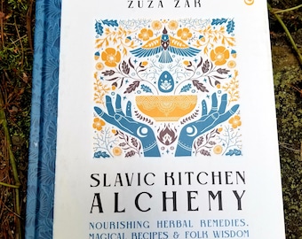 May include: A white book with a blue spine titled "Slavic Kitchen Alchemy: Nourishing Herbal Remedies, Magical Recipes & Folk Wisdom" by Zuza Zak. The cover features a colorful illustration of a bird, hands holding a bowl, and floral designs.