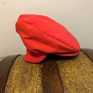 Puede incluir: Una gorra plana de color rojo brillante con una corona redondeada y una visera corta y rígida. La gorra tiene una banda de punto acanalado en la base. El sombrero está hecho de un material liso. La gorra está sobre un cofre de madera decorativo.