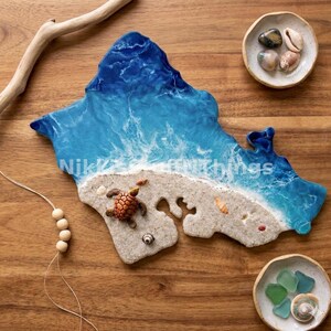 Puede incluir: Una pieza de arte de resina con forma de isla, que representa una escena de playa con un océano azul y una orilla arenosa. Una pequeña tortuga y conchas marinas están en la playa. El texto "Nikki's Things" es visible. También hay dos cuencos pequeños con conchas y vidrio marino.