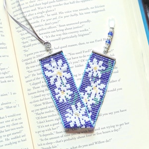 May include: A beaded bookmark featuring a blue and white snowflake design. The bookmark is crafted from small beads and has a silver chain and bead details at the top. It rests on the open pages of a book.
