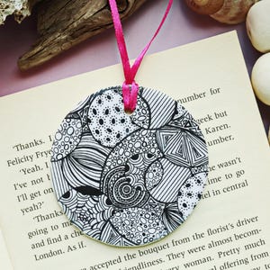 May include: A round, black and white patterned bookmark with a pink ribbon. The bookmark features a detailed, abstract design with various shapes and patterns. The bookmark is placed on an open book.