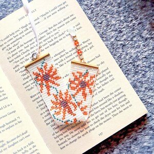 May include: A rectangular woven bookmark featuring a white background with orange and teal floral patterns, finished with gold-tone metal end caps. A white ribbon and a beaded strand with orange and clear beads are attached to the top of the bookmark.