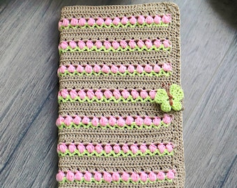 May include: A rectangular crochet notebook cover in a tan yarn base, featuring horizontal rows of light green and pink textured stitches. A small, light green crocheted butterfly is attached to the right side of the cover as a decorative accent.