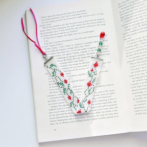 Könnte beinhalten: Handgemachter Lesezeichen mit weißer Basis, mit einem sich wiederholenden Muster aus roten Blumen und grünen Blättern. Das Lesezeichen hat ein rotes Band und silberne Metallenden. Es liegt auf einem Buch.