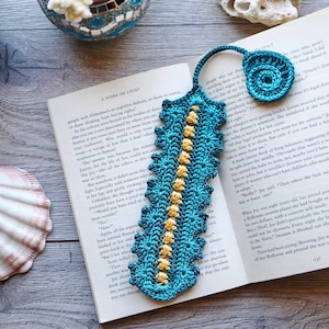 Op de afbeelding: Handgemaakte turquoise gehaakte bladwijzer met een spiraalvormig ontwerp en gele bloemen. De bladwijzer ligt op een open boek met de titel "A Spark of Light".